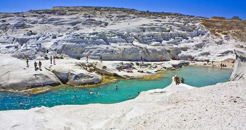 Thumbnail of Swimmers and sunbathers enjoy the turquoise water at Sarakiniko beach, surrounded by unique white volcanic rock formations.