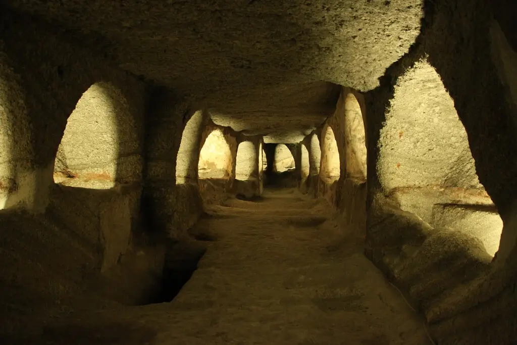 Thumbnail of A dimly lit, hand-carved underground tunnel of the Catacombs, showing arched burial niches along the walls.