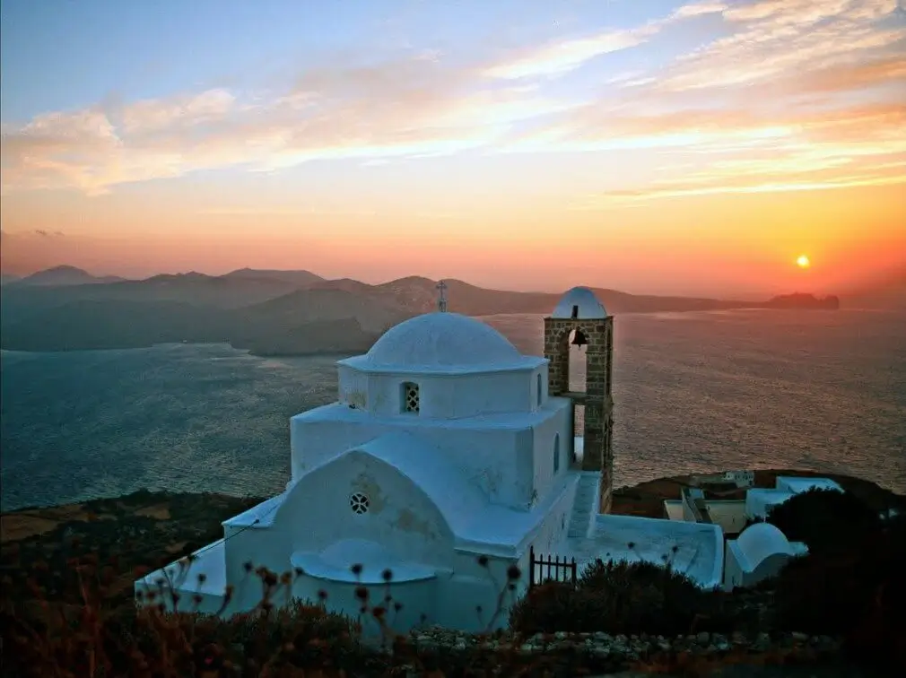 Thumbnail of A classic whitewashed Greek church with a dome, perched on a cliff overlooking the sea and islands at sunset.